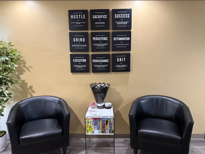 Waiting area with two black leather chairs facing a small table with a basket of magazines, beneath nine framed motivational posters reading Hustle, Sacrifice, Success, Grind, Persistence, Determination, Execution, Perseverance, and Grit